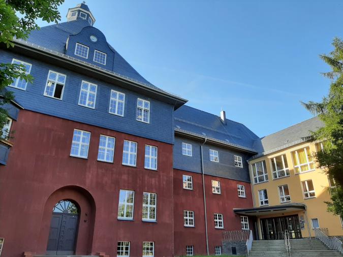 Blick auf das rot-gelbe Schulgebäude, umrahmt von zwei Bäumen, blauer Himmel