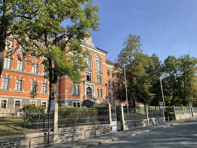 Blick auf das Backsteinschulgebäude von der Straße aus, mit Zaun und grünen Bäumen